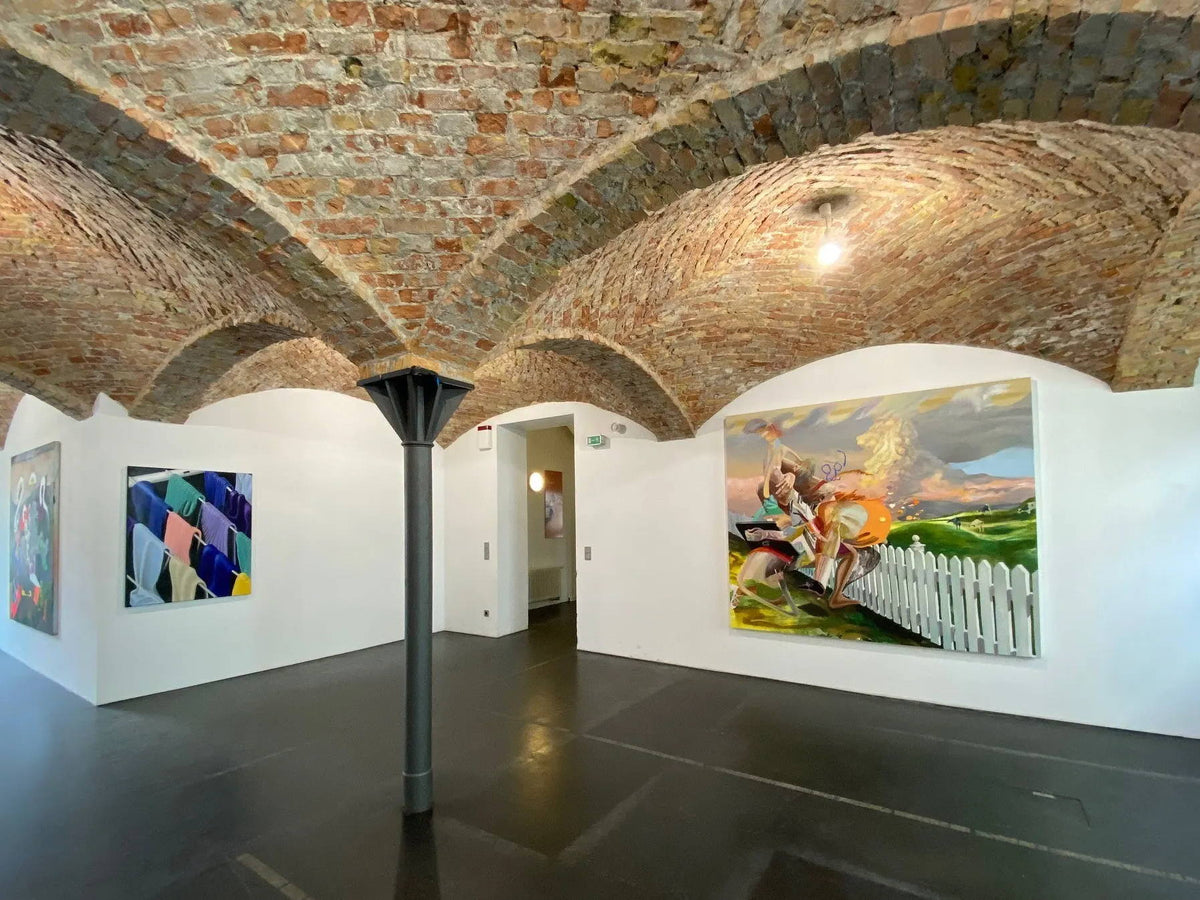Galerie d'art avec un plafond voûté en briques et des tableaux aux couleurs vives accrochés sur des murs blancs.