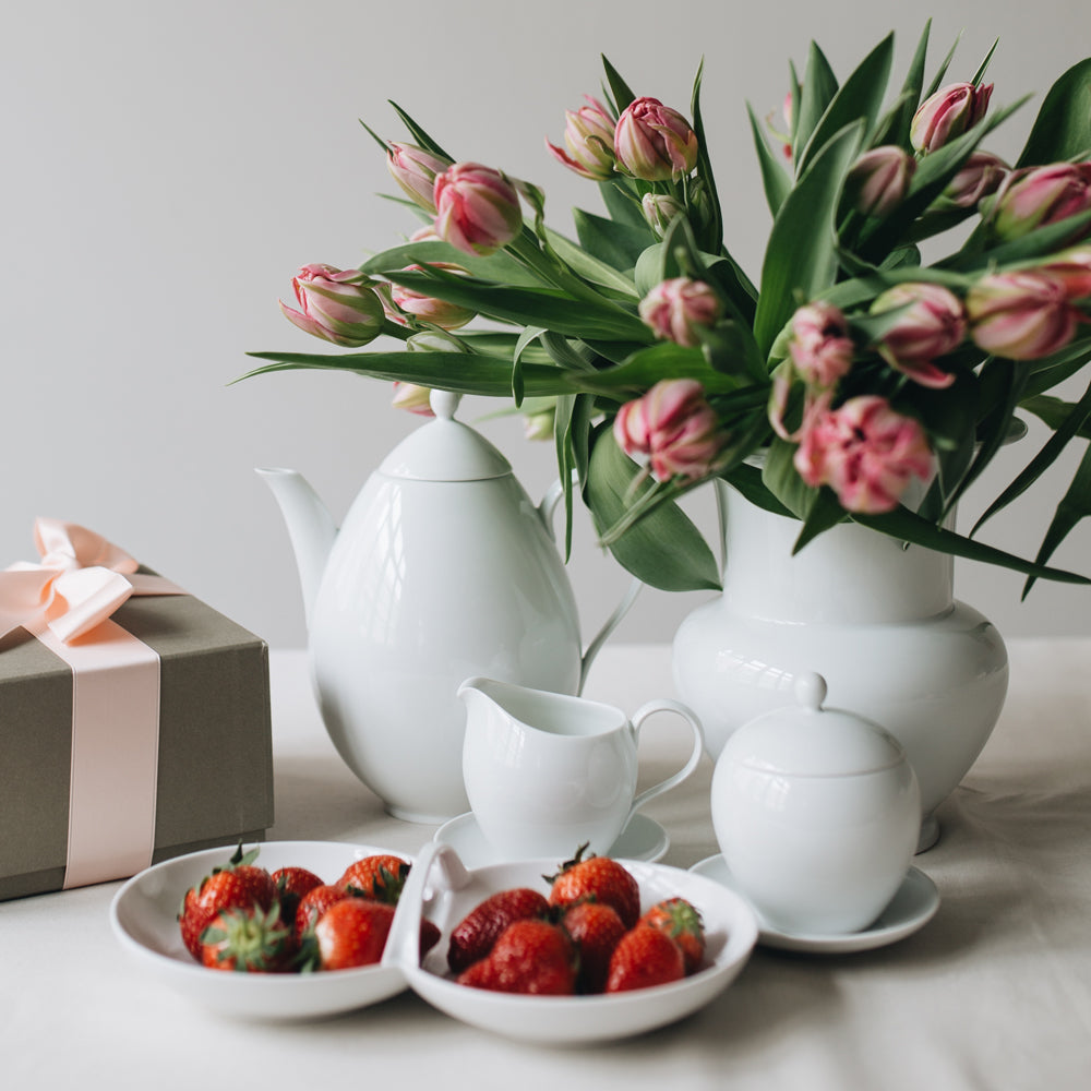 Composition florale avec une théière, un pot à crème et des fraises sur une table.