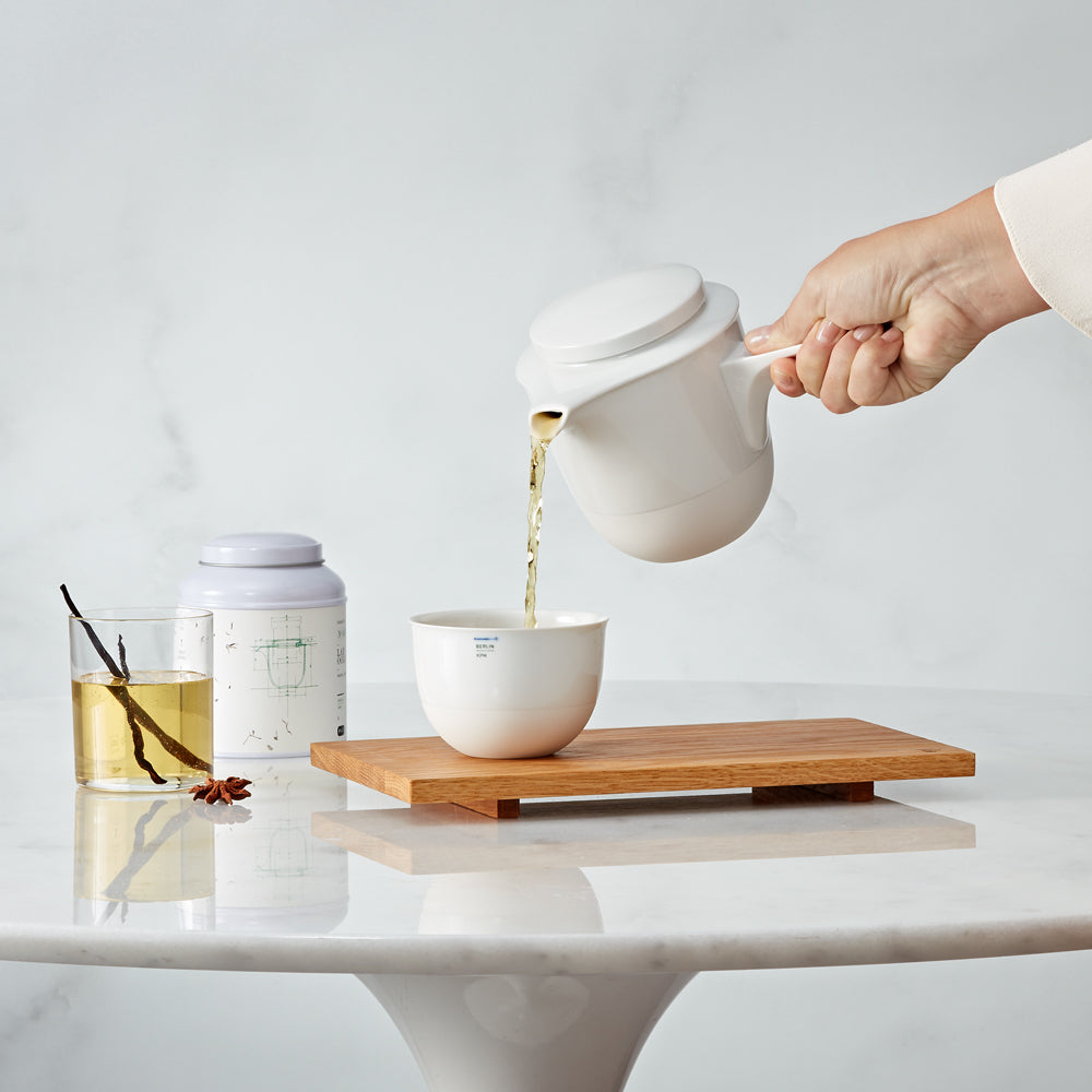 Une personne verse du thé depuis une théière blanche KPM LAB dans une tasse posée sur une table.