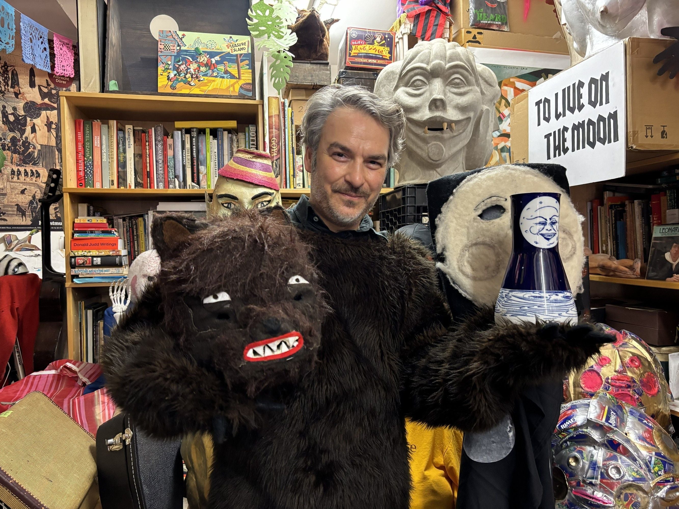 Person in a bear costume surrounded by various masks and bookshelves.